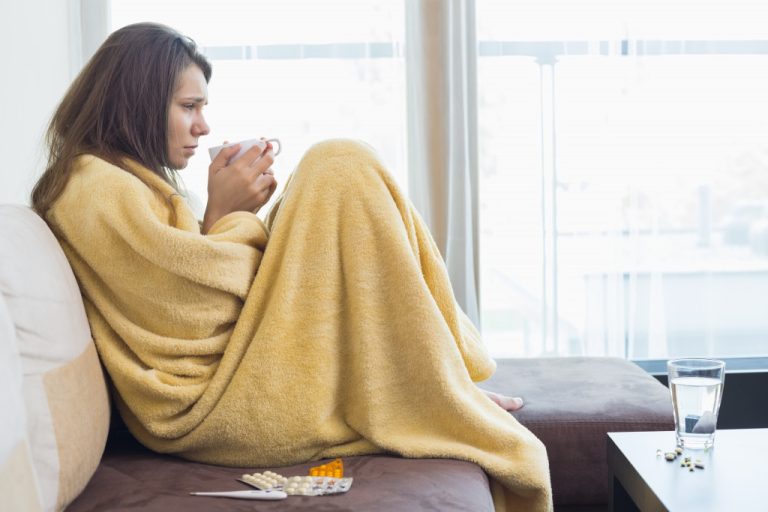 A sick woman wrapped in a blanket drinking coffee and medicine on a living room sofa