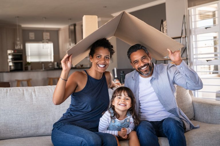 A family sitting in the new home while holding a cardboard to mimic a roof