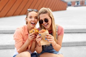 Two young womne smiling while eating burgers in the city.