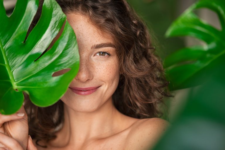 a curly haired woman with freckles smiling while hiding behind leaves