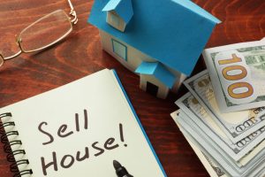 Sell House! written on a notebook, a house, glasses, and money on a table